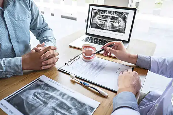 Dentist explaining full-mouth restoration treatment to a patient using X-rays, a teeth model, and notes on a desk.